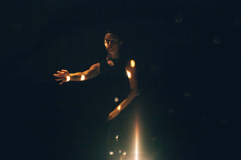 Rachel Mckinstry in a black tank top broods with her arms outstretched. A light shines right on her.